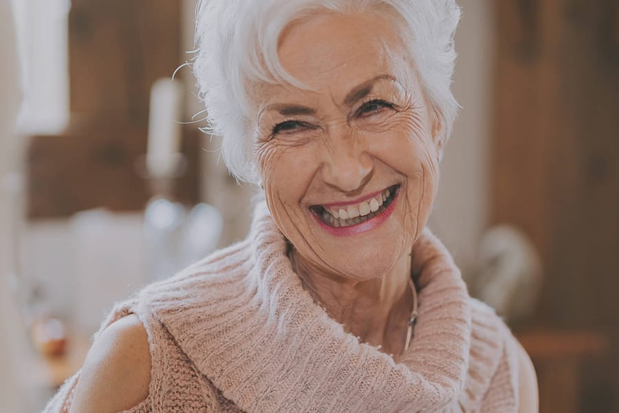 Smiling older woman with short white hair, wearing a soft pink knitted jumper, looking warmly toward the camera in a cozy indoor setting.
