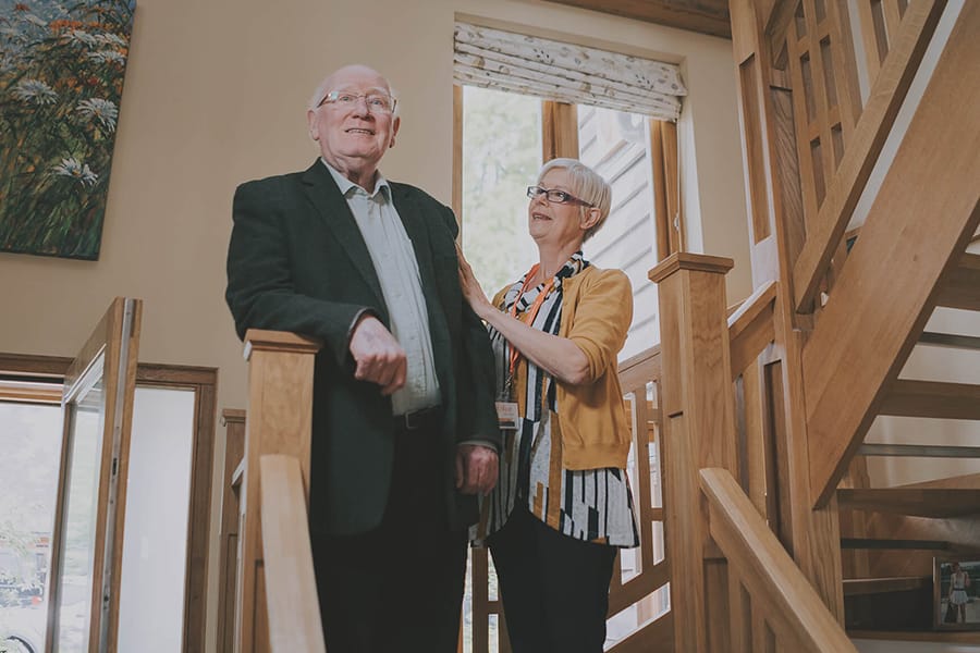 An older man and woman standing together on a wooden staircase in a bright home. The woman is gently touching the man’s arm as they smile and look ahead, surrounded by natural wood features and large windows.