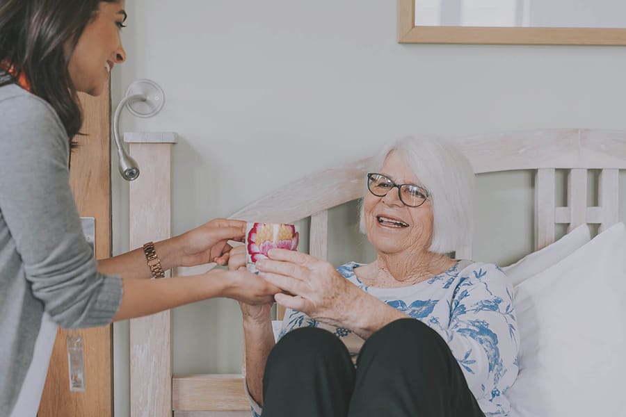 An older woman sitting up in bed and accepting a colourful mug from a younger woman carer who is standing beside the bed. The older woman is smiling, and the scene takes place in a bright bedroom with light-coloured furniture.