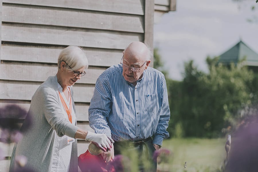 An older man and an woman carer standing outside near a wooden building, looking down at plants or flowers. The woman is wearing gloves and is tending to the garden while the man observes beside her.