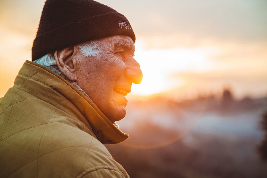An elderly man wearing a beanie and a warm jacket smiles while looking into the sunset outdoors