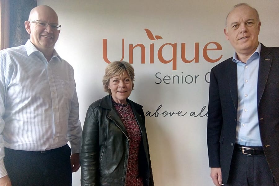 Three adults standing in front of a wall with the logo “Unique Senior Care,” smiling toward the camera