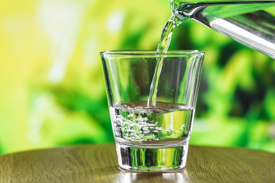 Clear water being poured from a glass pitcher into a transparent glass on a wooden surface, with a bright green blurred background