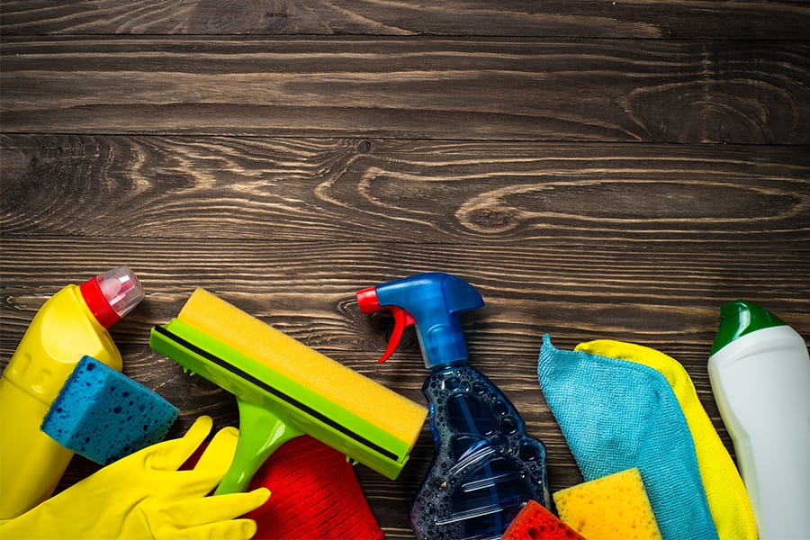 A collection of cleaning supplies, including spray bottles, sponges, rubber gloves, and cloths, arranged along the bottom edge of a wooden surface