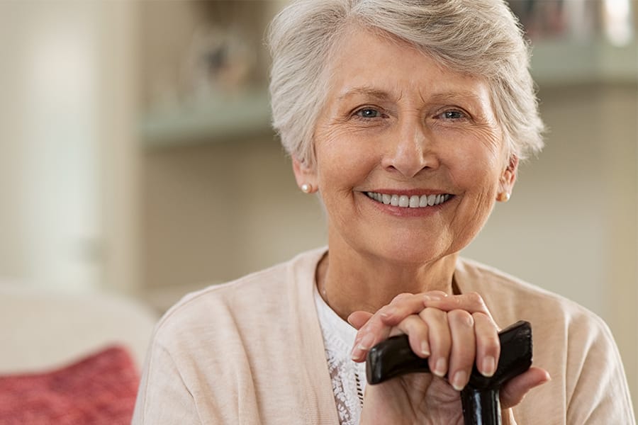 An elderly woman with short gray hair smiles warmly while resting her hands on the handle of a cane