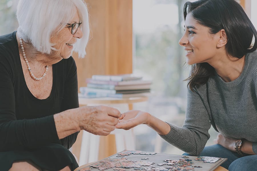 An elderly woman and a younger woman smile warmly at each other while working on a jigsaw puzzle together at a table.