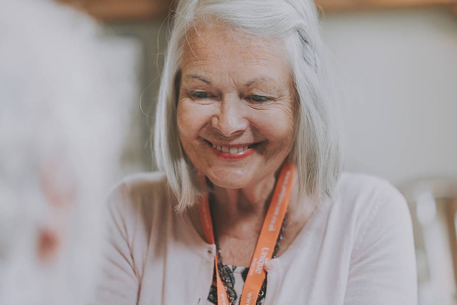 A woman carer with gray hair smiles warmly while wearing a lanyard and light-colored clothing.