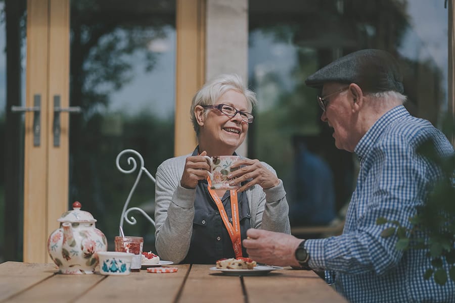 An elderly woman carer smiles while having tea with an elderly man at an outdoor wooden table, with cups, pastries, and a teapot in front of them.