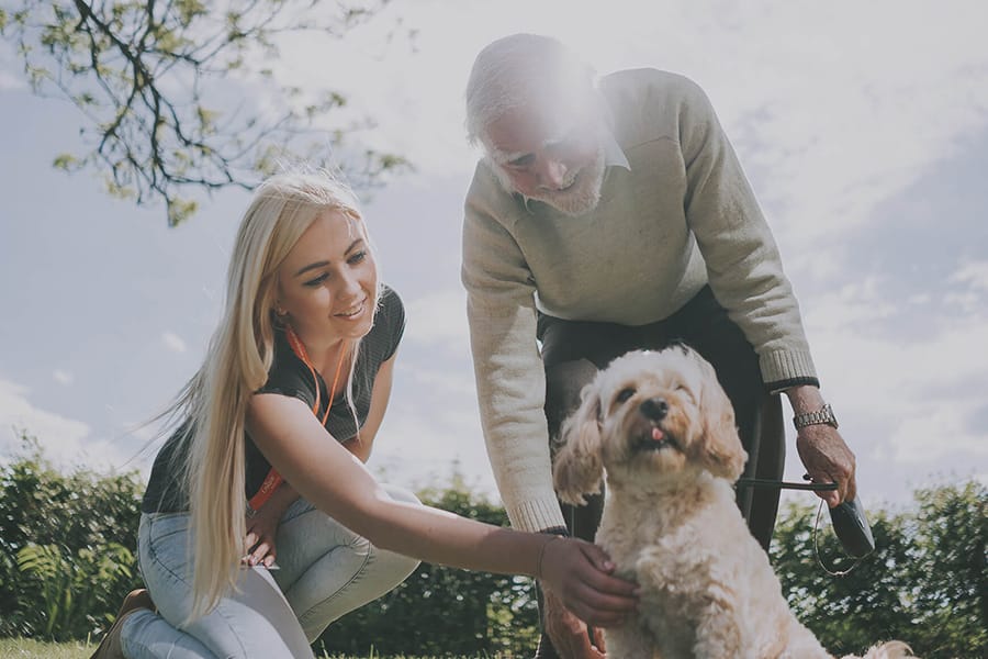 A carer kneels beside an elderly man as they both smile and pet a small dog outdoors on a sunny day