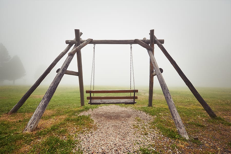 An empty wooden swing hangs from a rustic frame in a foggy outdoor field, creating a quiet and atmospheric scene