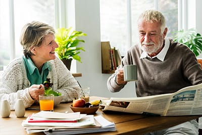 An elderly couple sits at a kitchen table enjoying breakfast together, with the man reading a newspaper and holding a mug while the woman smiles and eats fruit.