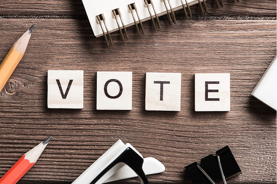 Wooden letter tiles spelling out “VOTE” arranged on a desk surrounded by pencils, clips, and office supplies.