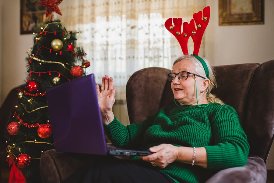An elderly woman wearing festive reindeer antlers sits in an armchair beside a decorated Christmas tree, waving at a laptop.