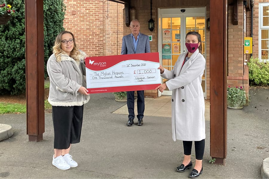 Two women hold a large charity cheque for £1,000 made out to The Myton Hospices, while a man stands behind them outside a building. One woman is wearing a face mask.