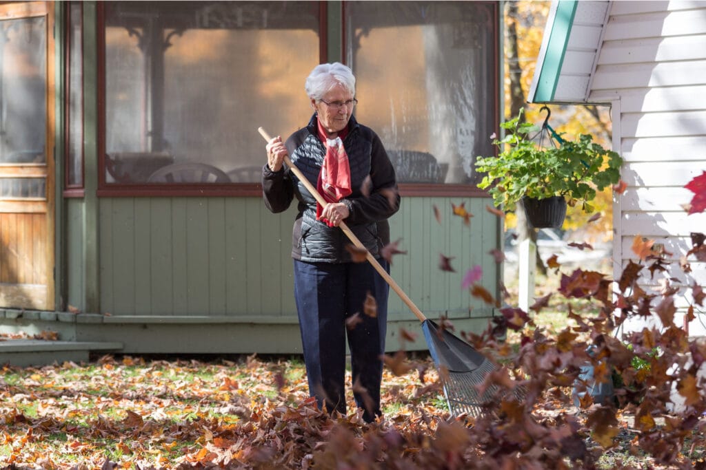 An elderly woman rakes fallen autumn leaves in her yard, standing near a house with a hanging plant and wearing a scarf and jacket