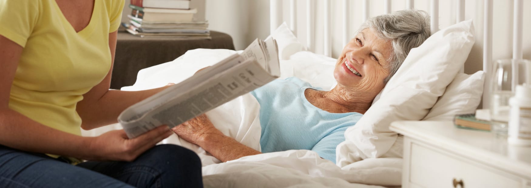 elderly woman in bed, with a carer sat reading