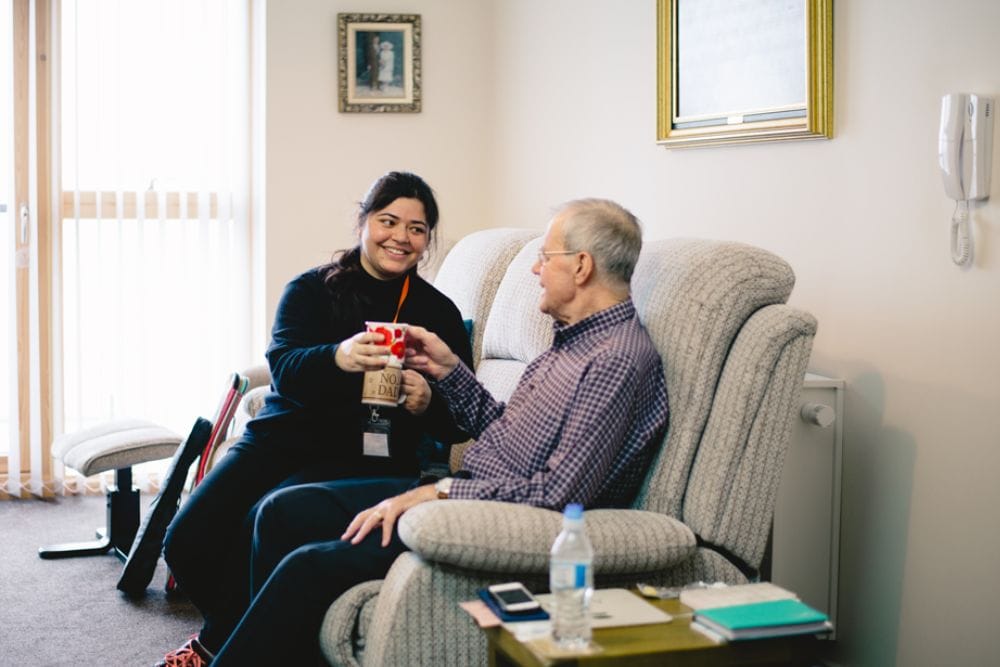 A Unique Senior Care employee with an orange lanyard around her neck, sits sharing a cup of tea with an elderly live in care companion.