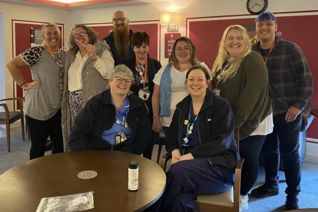 A group of care workers and staff stand and sit together in a brightly lit room, smiling for the camera. Two people in NHS uniforms are seated at the front, while the rest of the team stands behind them. A round table is in the foreground with a bottle and some items on it.