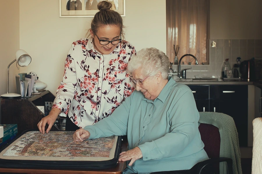 An elderly woman and her caregiver work on a large puzzle together at a table in a well-lit room, emphasizing the factors that cause live-in care costs to vary in Somerset.