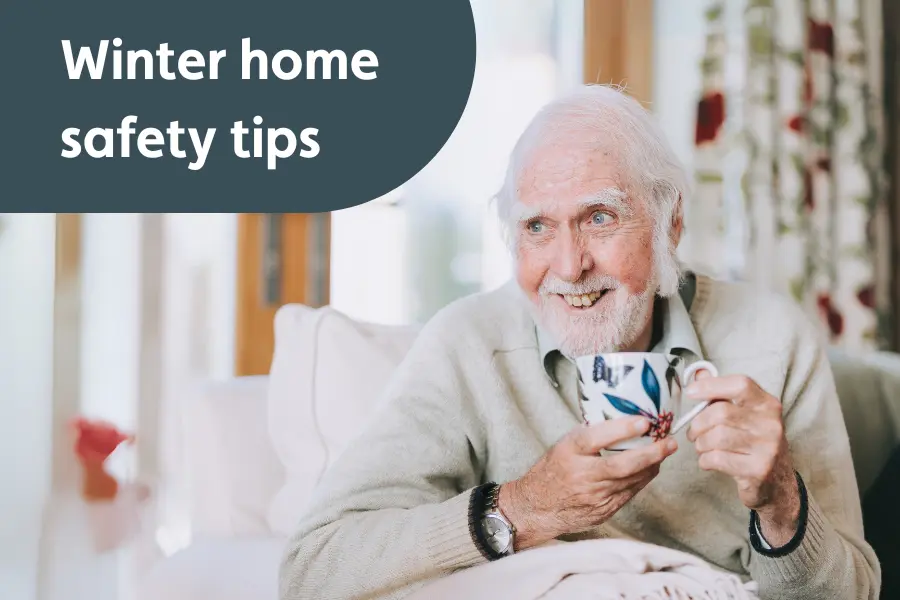 An elderly man with white hair and a beard sits indoors, smiling warmly while holding a patterned mug with both hands. He is covered with a light blanket and appears relaxed. In the upper left corner, a dark grey rounded box contains the text “Winter home safety tips.”