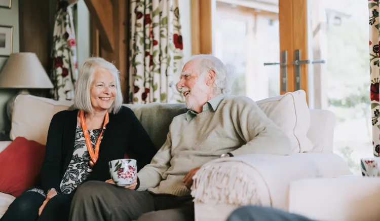 Picture of lady carer sitting on the sofa with elderly gentleman client smiling and chatting together
