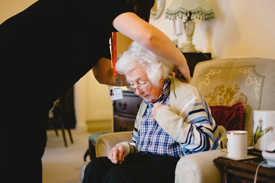 A caregiver assisting an elderly woman seated in a beige floral armchair. The caregiver adjusts the woman’s cardigan in a warmly lit living room, with a cup of tea on a nearby table. Displayed in the ensuring compatibility section of the Merseyside and Wirral Live-In Care Page.
