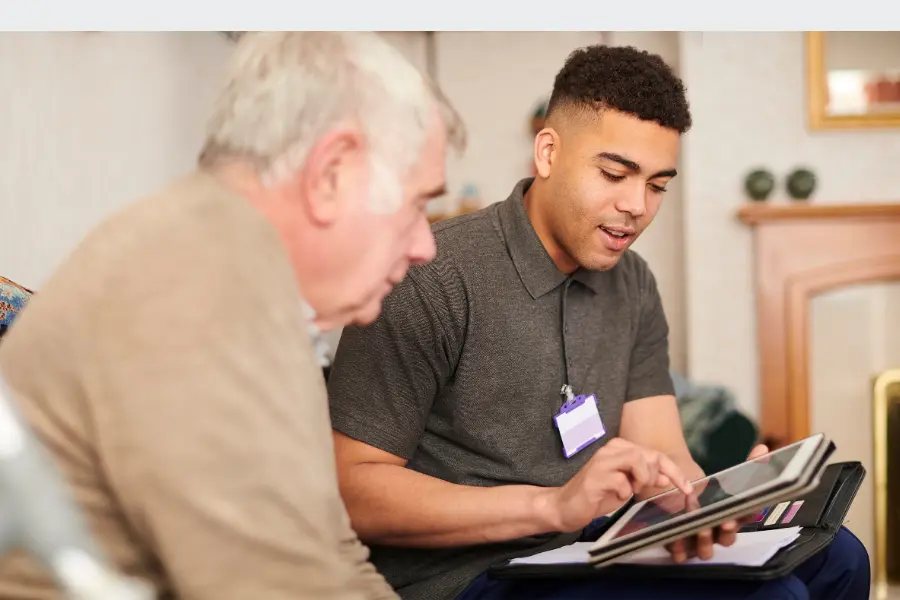 A caregiver sitting with an older man, using a tablet to review or explain information in a home setting.