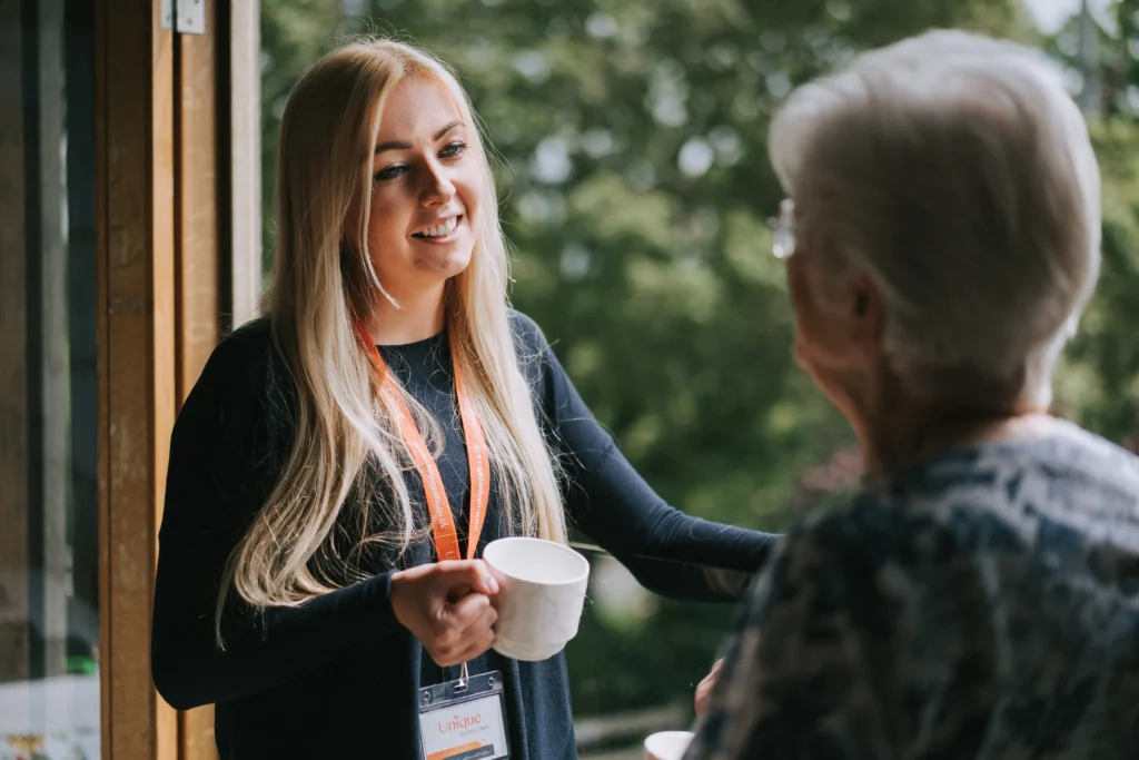 A caregiver with a lanyard smiling warmly while holding a mug, chatting with an elderly woman in a relaxed, scenic setting.