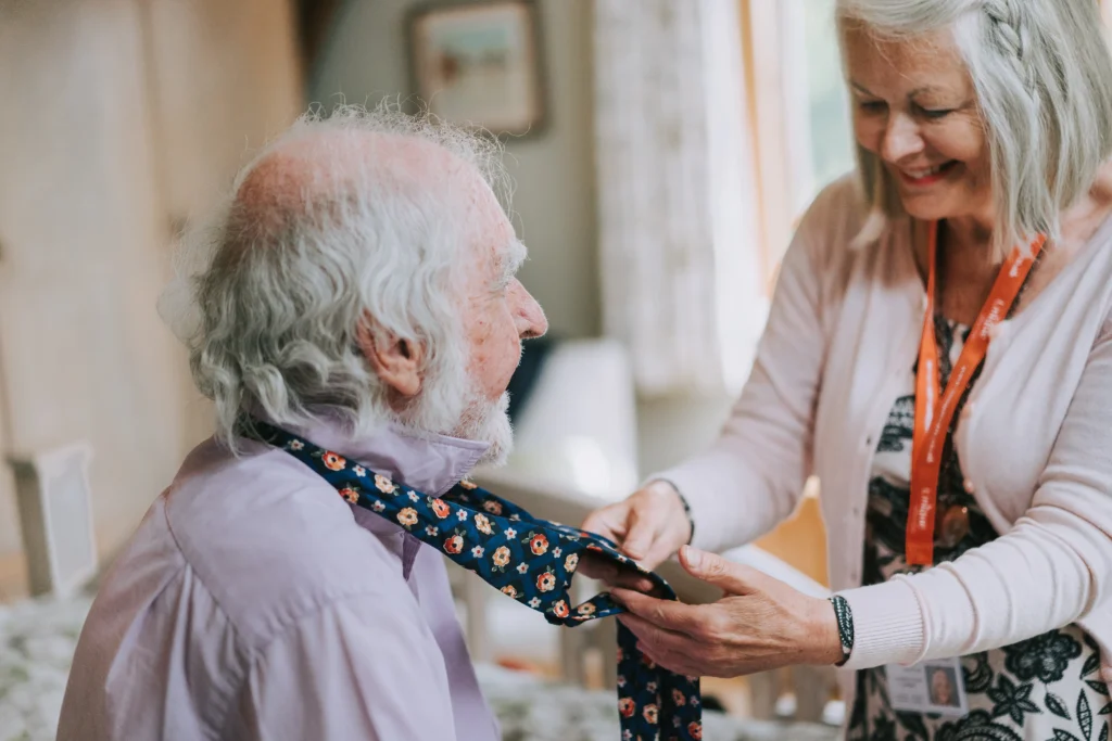 A caregiver helping an elderly man adjust his floral tie, smiling in a warm home setting.