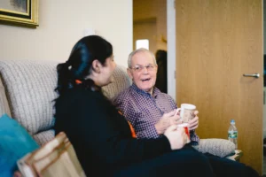 A caregiver sitting on a sofa, smiling and chatting with an elderly man while they both hold mugs in a cosy living room.