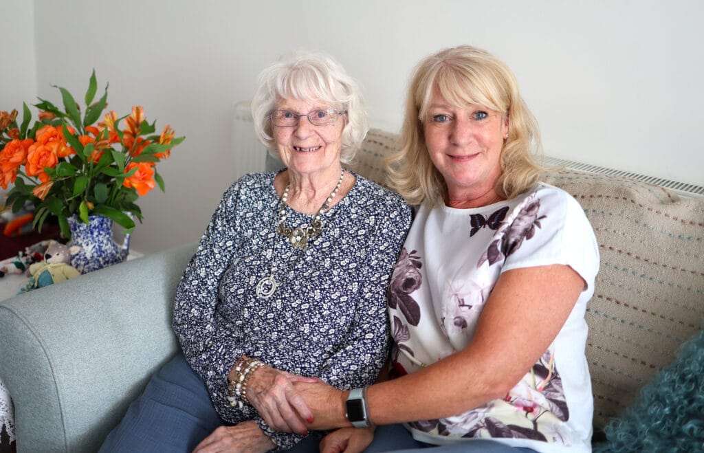 An elderly woman and a middle-aged woman sit closely together on a sofa, smiling warmly at the camera. The elderly woman has white hair, glasses, and wears a patterned blue top with layered necklaces. The other woman has blonde hair and wears a white floral top, gently holding the elderly woman’s hand. A vase of orange flowers sits to the side, adding colour to the scene.