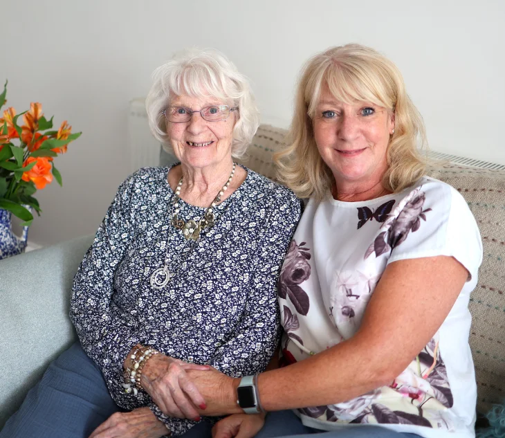 An elderly woman and a middle-aged woman sit closely together on a sofa, smiling warmly at the camera. The elderly woman has white hair, glasses, and wears a patterned blue top with layered necklaces. The other woman has blonde hair and wears a white floral top, gently holding the elderly woman’s hand. A vase of orange flowers sits to the side, adding colour to the scene.