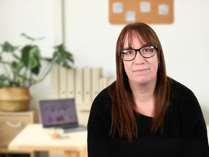 A woman with long brown hair and glasses sits indoors, looking toward the camera with a neutral expression. She is wearing a black top. Behind her is a softly blurred office setting with a desk, an open laptop, folders on a shelf, a potted plant, and a noticeboard on the wall.