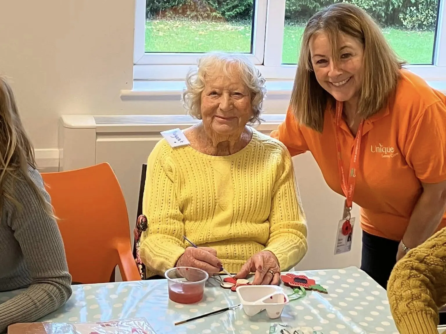 An older woman in a bright yellow jumper enjoys a painting activity at the Memory Café, sitting beside a staff member in an orange Unique Senior Care shirt.