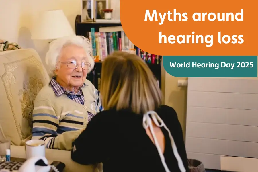 An elderly woman sitting in an armchair, smiling while talking to a caregiver who is facing her. It's overlayed with the test 'Myths around hearing loss. World Hearing Day 2025)