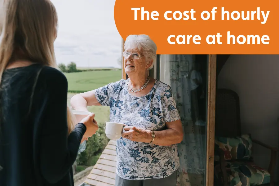An elderly lady, stood by a window smiling at her caregiver while holding a cup of tea, overlayed with the text 'The cost of hourly care at home'