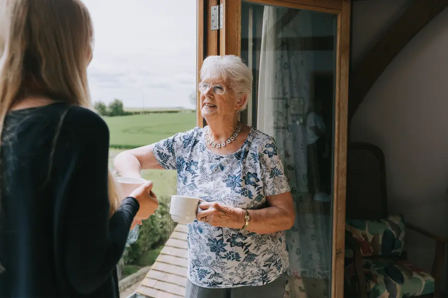 An older woman stood by a window smiling at her caregiver while holding a cup of tea