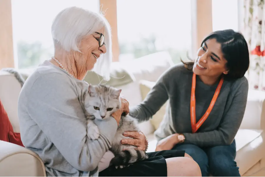 A caregiver and older lady on the sofa together smiling while stroking her cat