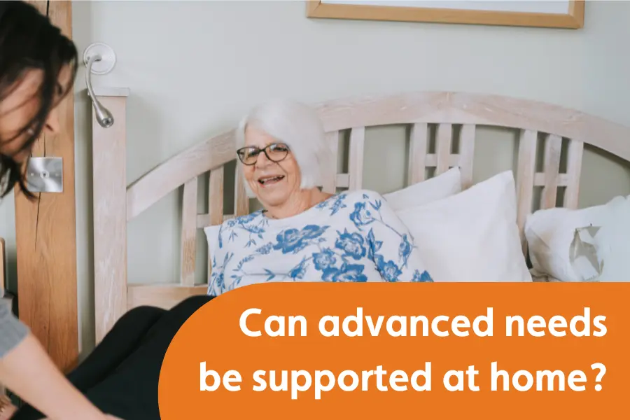 An elderly woman with white hair and glasses sits up in bed smiling, while a carer assists nearby. A large orange banner asks, “Can advanced needs be supported at home?”.