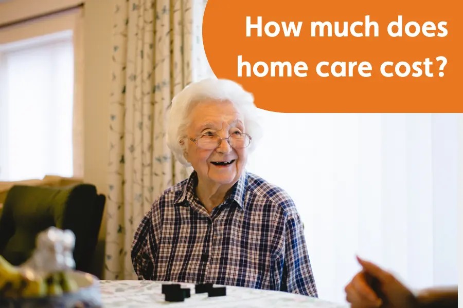 Thumbnail image for 'how much does home care cost' page, featuring an elderly lady sat at a table smiling at someobdy