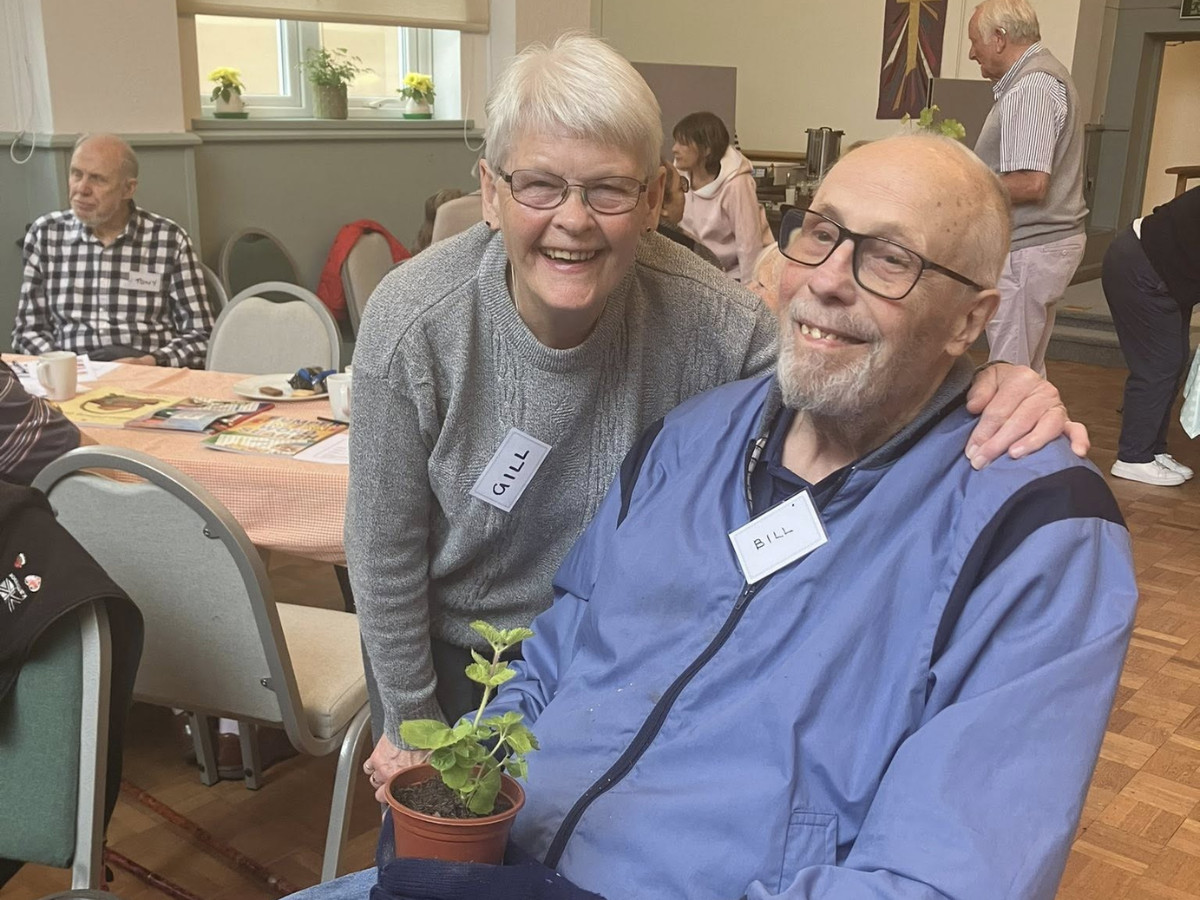 Two attendees, Gill and Bill smiling together at a memory café, with Bill holding a small potted plant.