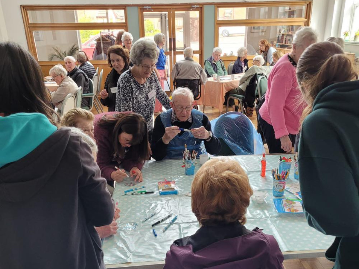 A busy table at a memory café, with older people and volunteers enjoying a craft activity together.