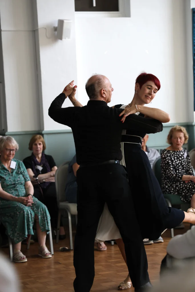 Professional dancers and attendees in a hall, with guests seated and clapping along during a performance at the Memory Café Tea Dance.