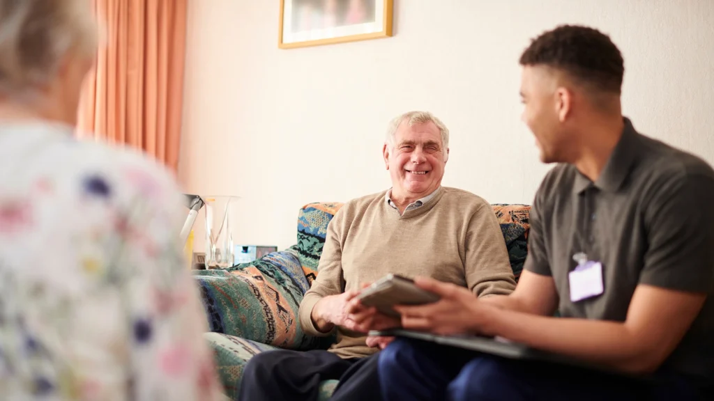 Older man smiling on the sofa while chatting to a care professional during a home visit