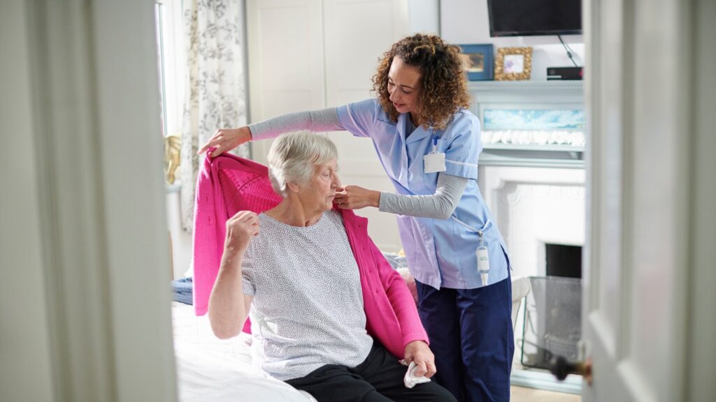 Caregiver helping an older woman put on a cardigan in a bright, comfortable bedroom