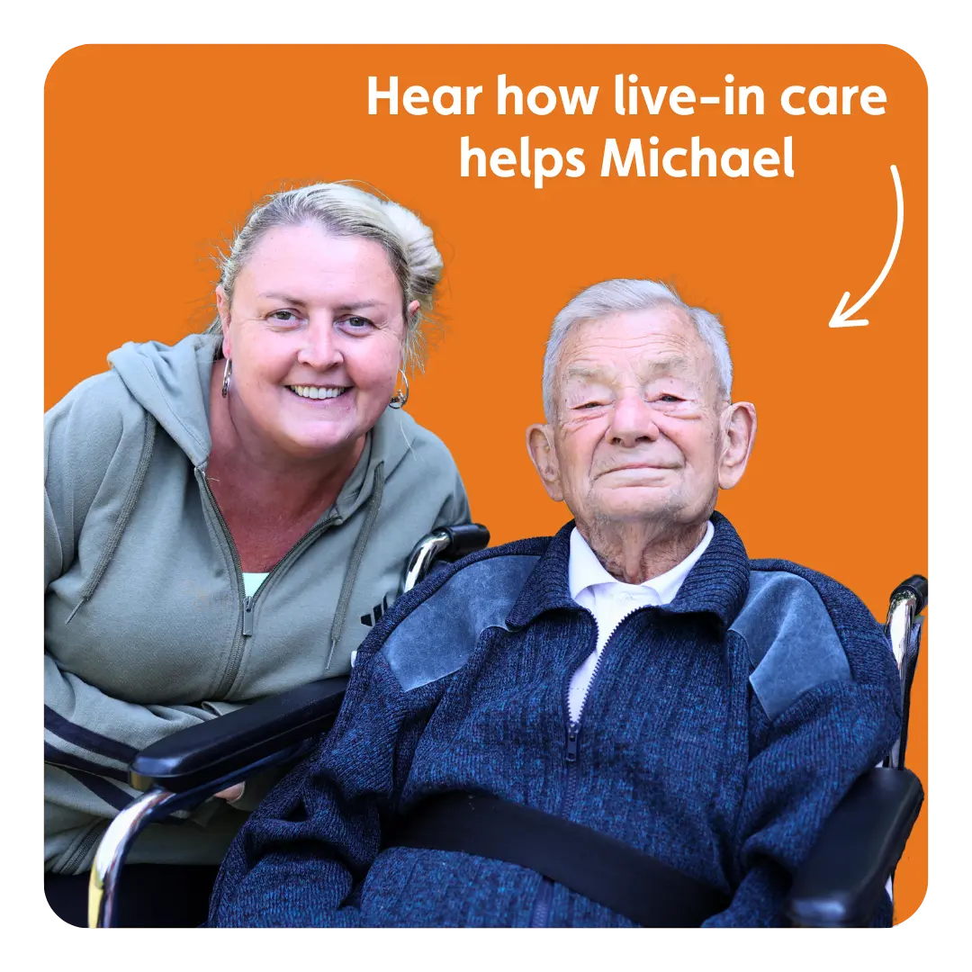 A smiling carer leans beside an elderly man seated in a wheelchair, with an orange background and text saying “Hear how live-in care helps Michael.”