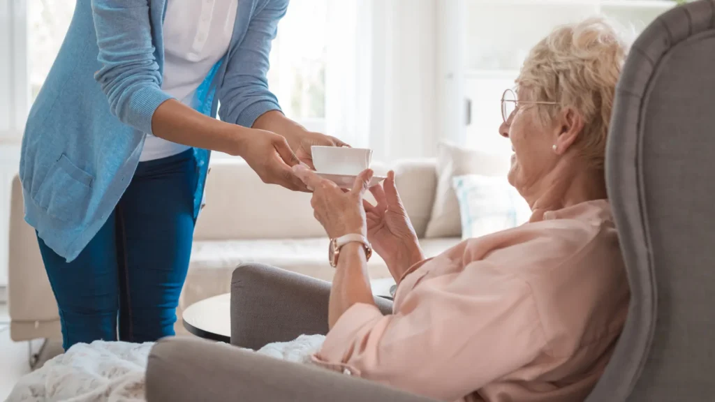 A lady handing a cup of tea over to a smiling older lady sat in a chair