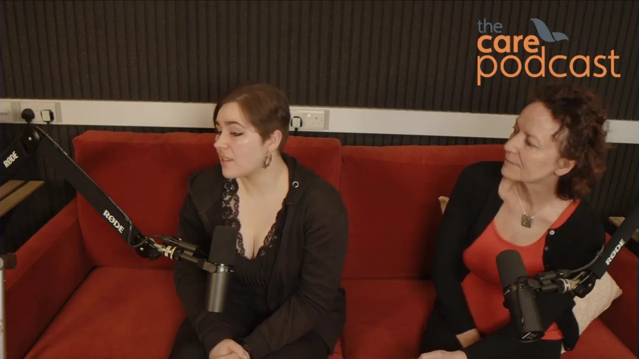 Two podcast guests on a red sofa with studio microphones, engaged in attentive conversation.