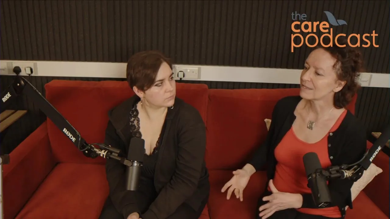 Mother speaking with hand gestures while daughter listens on a red sofa during a podcast interview with microphones