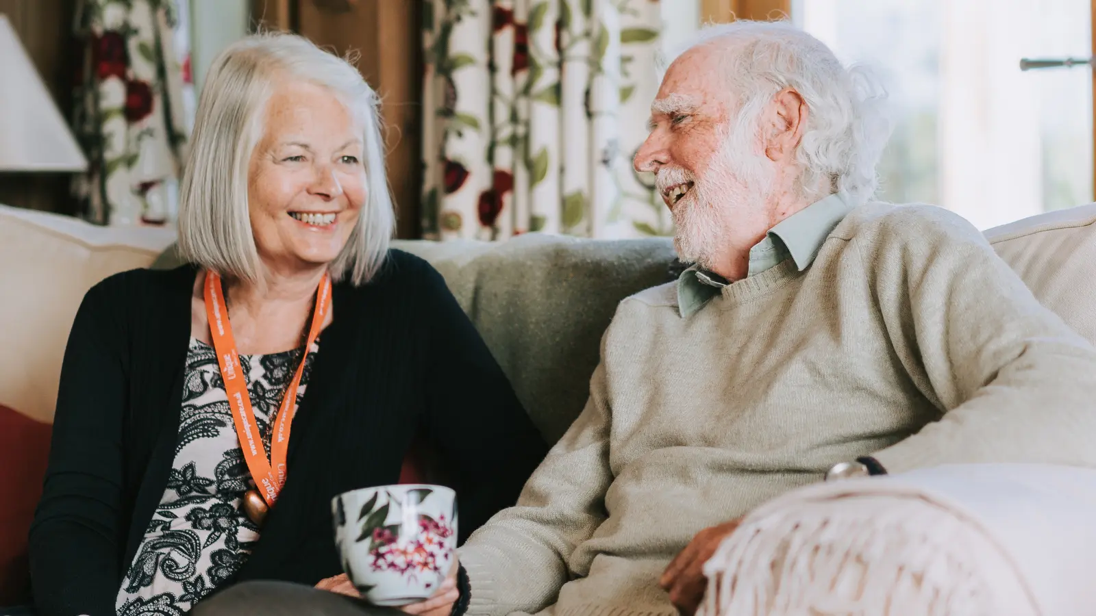 Picture of lady carer sitting on the sofa with elderly gentleman client smiling and chatting together